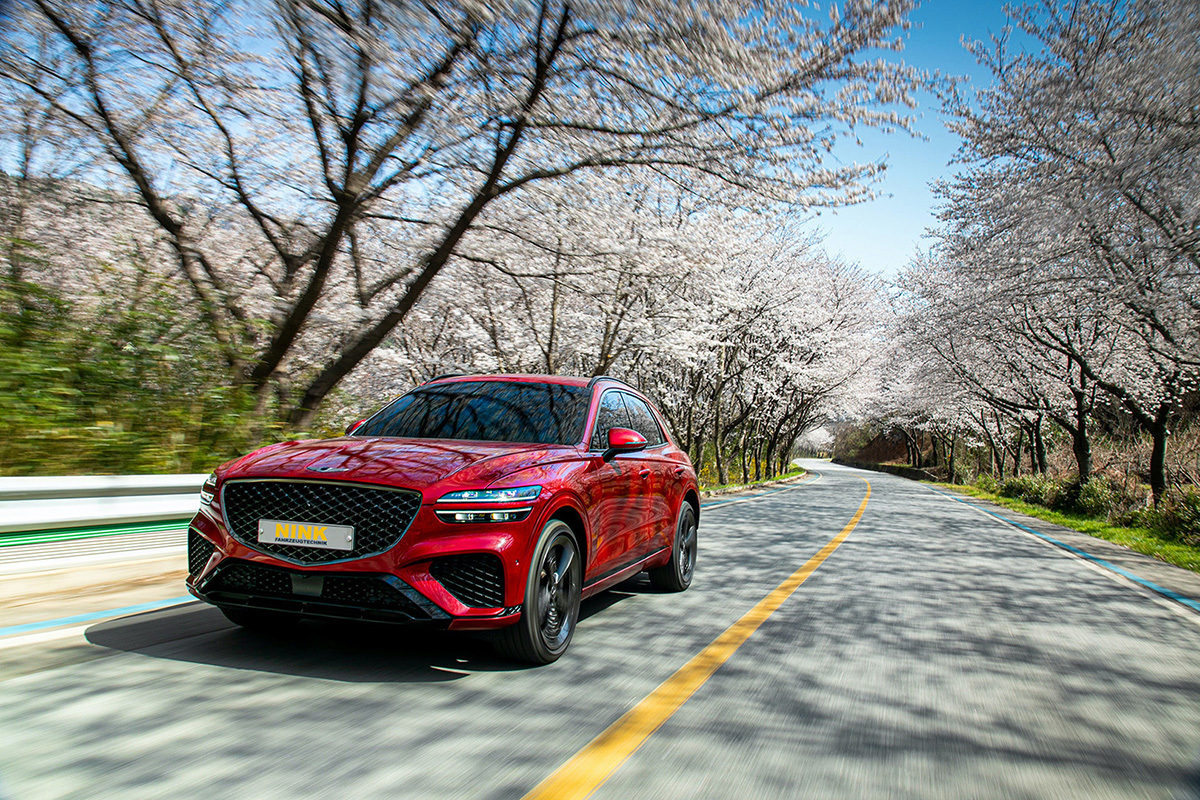 Red luxury SUV driving on a scenic road lined with blooming cherry trees, symbolizing a spring vehicle check from Fahrzeugtechnik Nink.